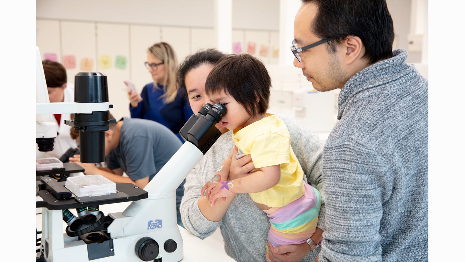 Parents of a toddler hold her while she looks into a microscope